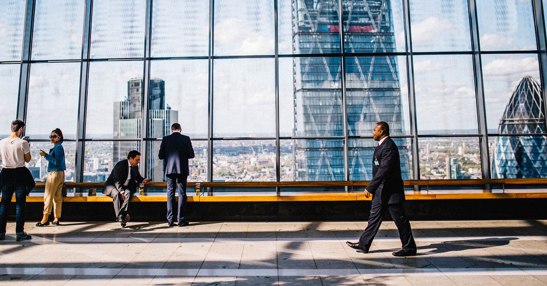 A man in a suit walks along a glass corridor. Skyscrapers are visible outside the windows.