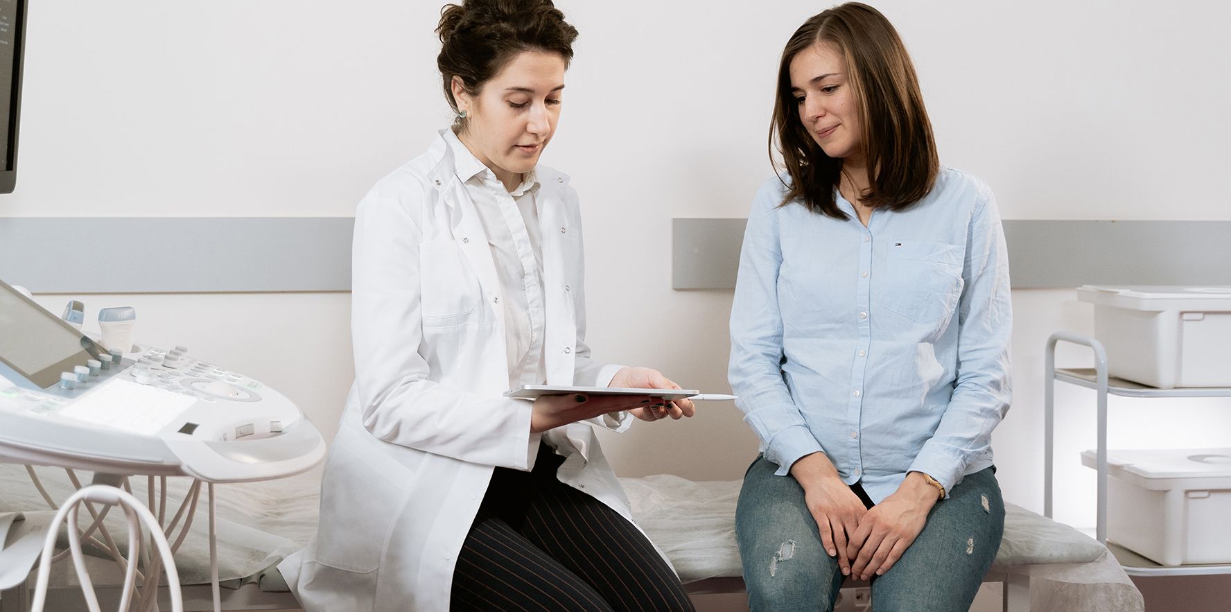 A female doctor in a lab coat talks with a female patient in an exam room.