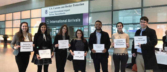 Seven students in business attire pose for a photo at Logan Airport. Each holds a sign that reads "legal help."