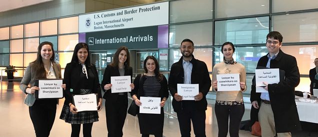 Seven students in business attire pose for a photo at Logan Airport. Each holds a sign that reads "legal help."