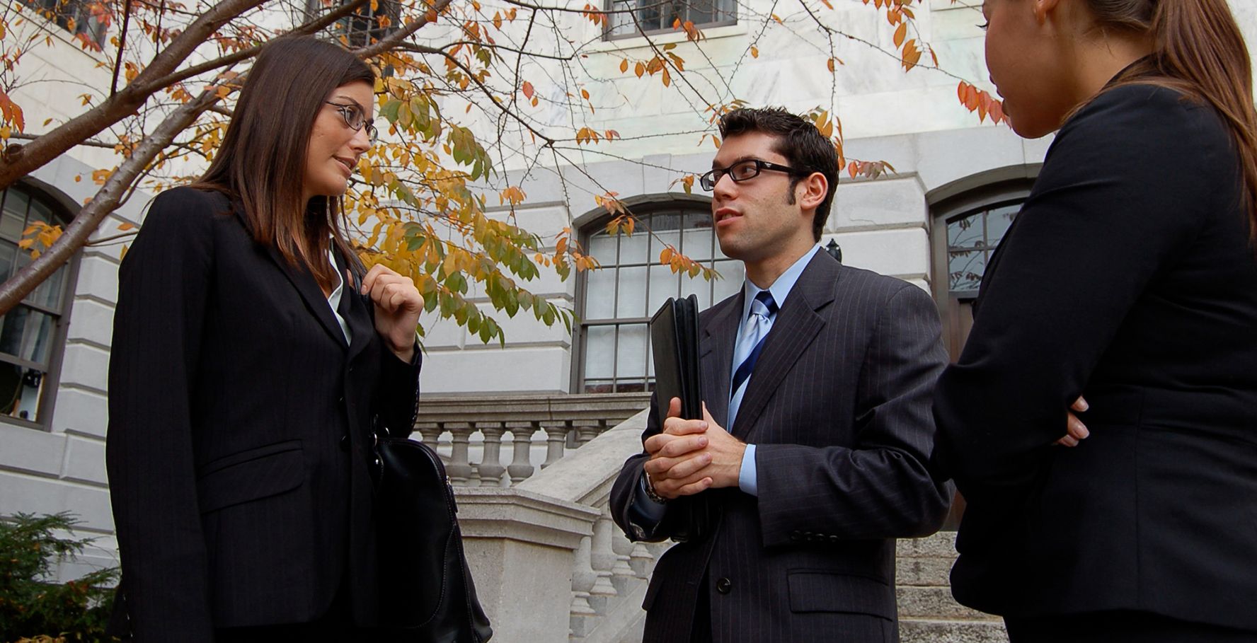 Three students in business attire stand outside a courthouse. They hold briefcases and folders.