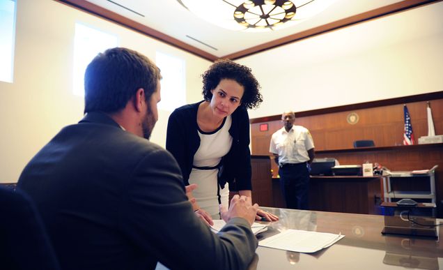 Two students discuss a case in a large courtroom. A bailiff stands in the background.