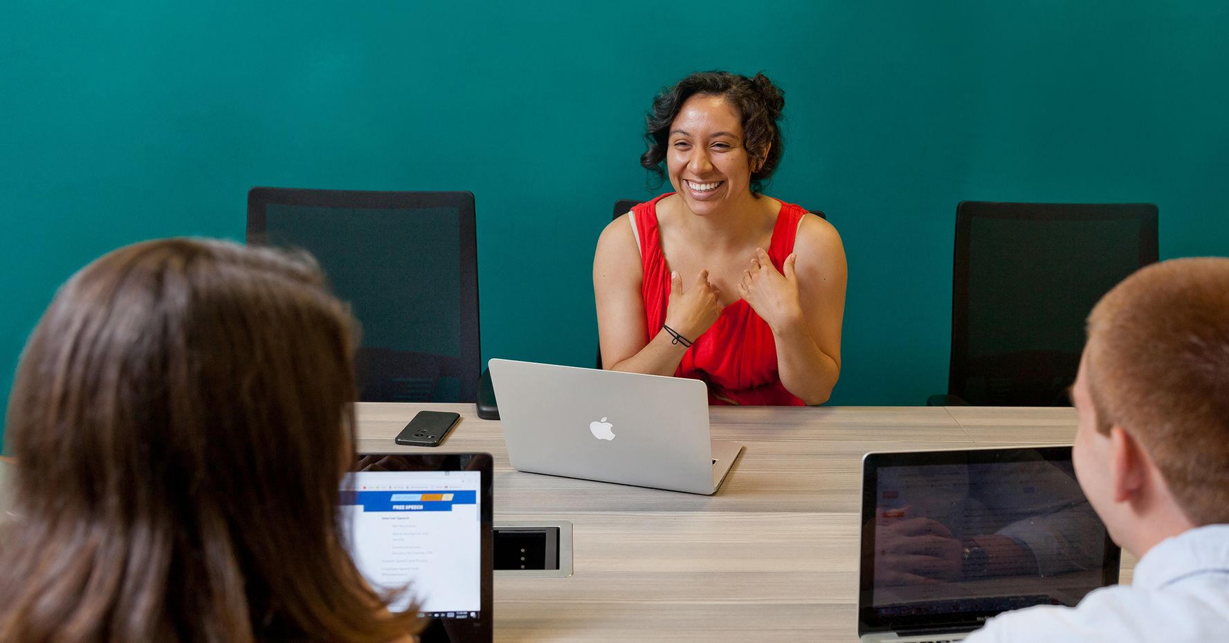 A smiling female student speaks to classmates on the other side of a conference table. All three have laptops open.