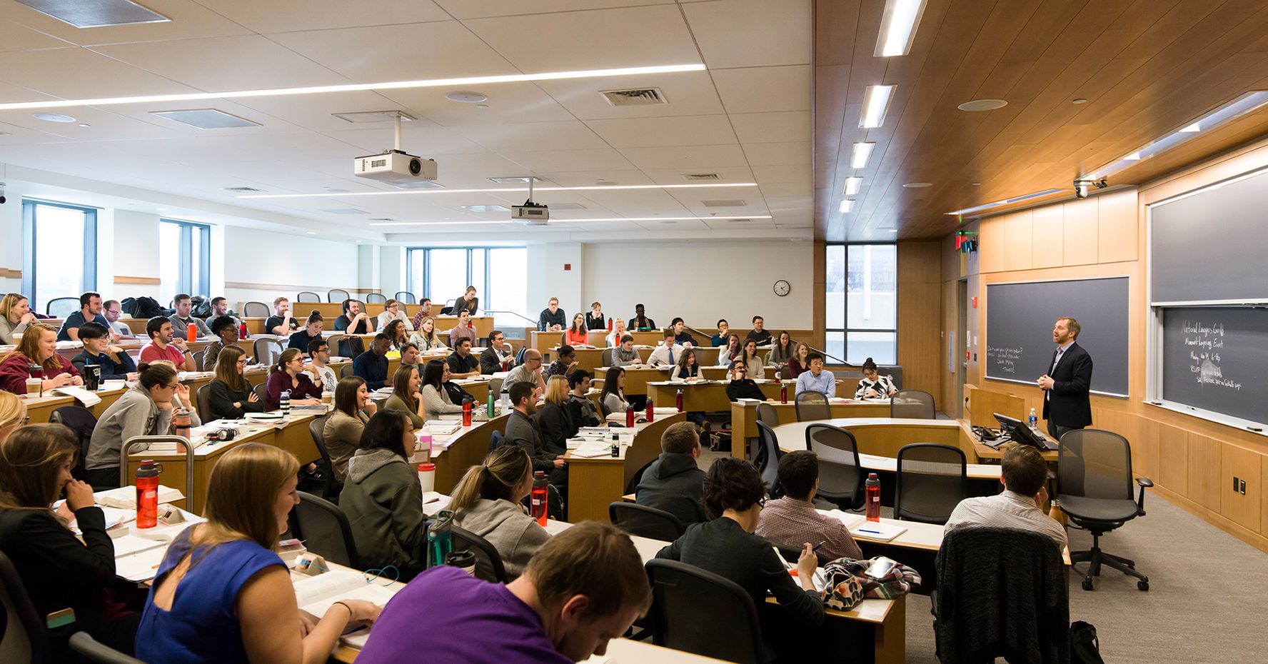 A professor stands against a blackboard and speaks to students in a full lecture hall.