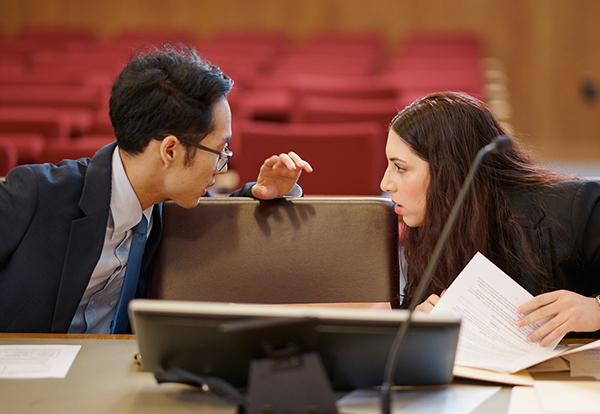 Two students lean toward each other and speak quietly in a courtroom.