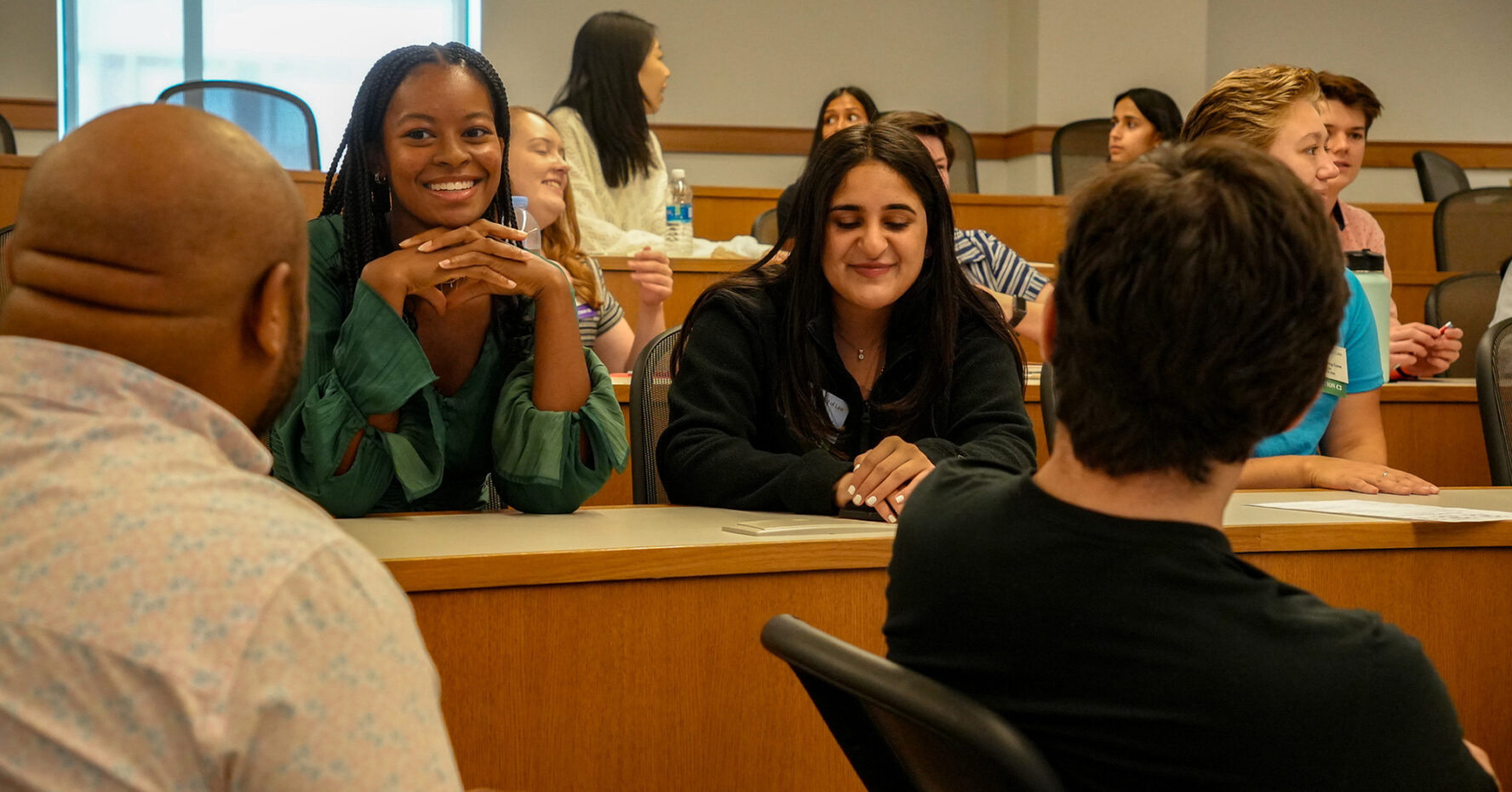Two smiling students sit in a lecture hall conversing with others.