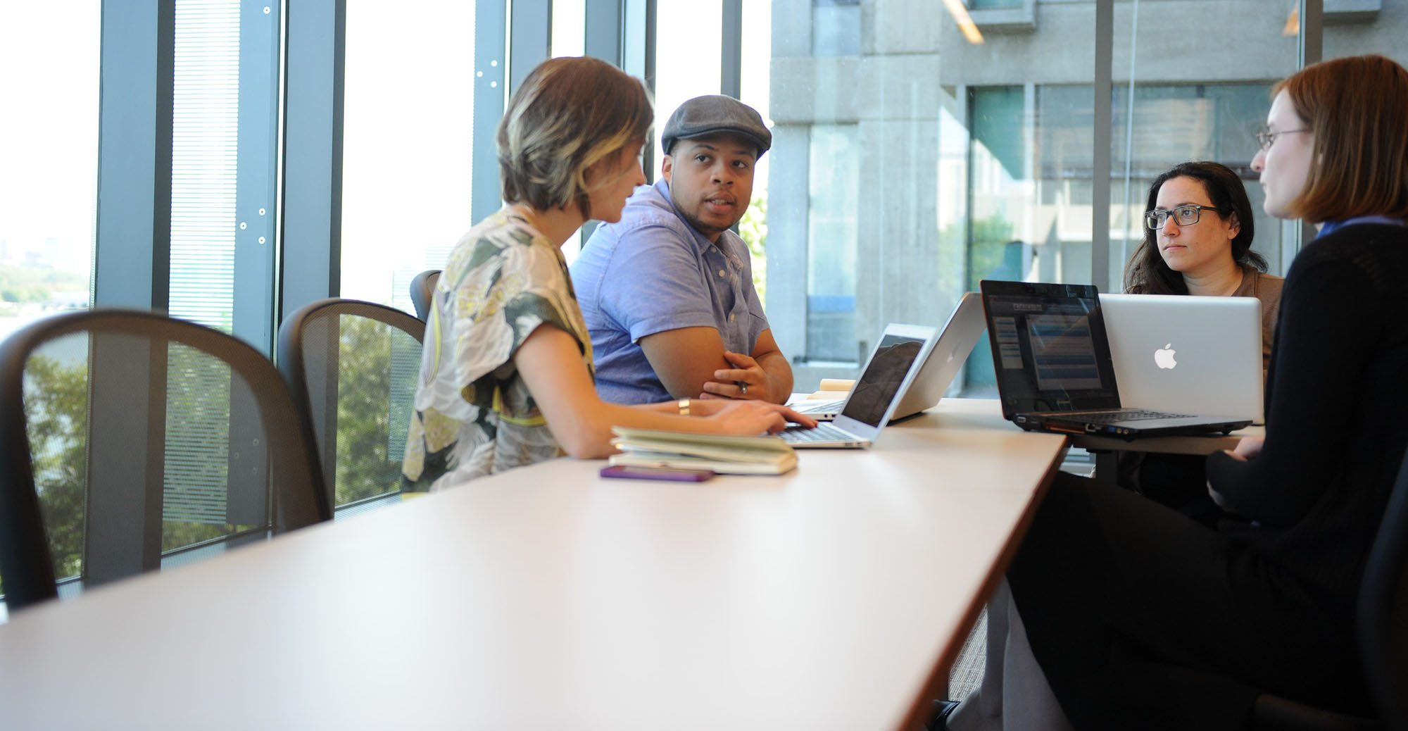 Students meet at a conference table in a large, bright classroom with many windows.