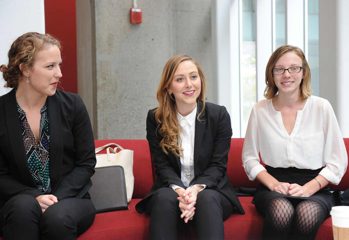 Three students in professional attire sit on red couches in the atrium of the law complex.