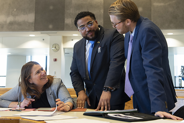 Two students dressed in suits speak with an instructor in a courtroom.