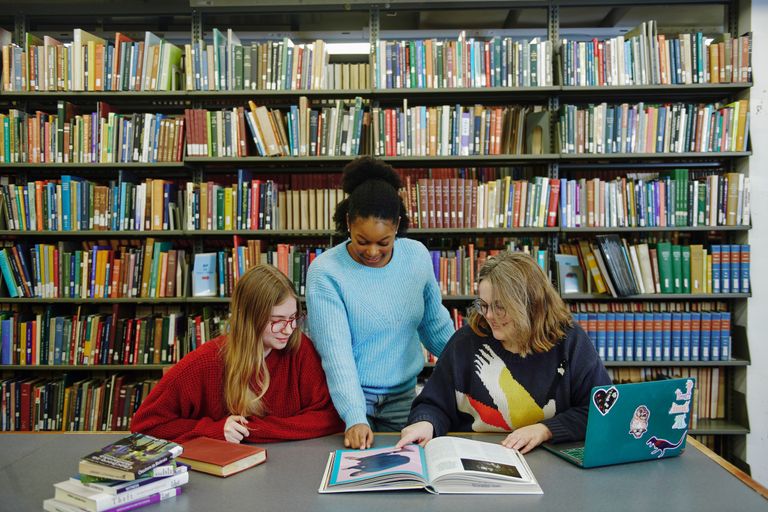 Two students sit at a desk looking at a book, while a third student stands between them pointing out content in the book.