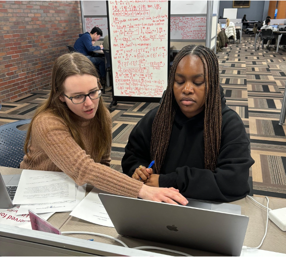 A student points at a laptop computer screen while another student looks on.