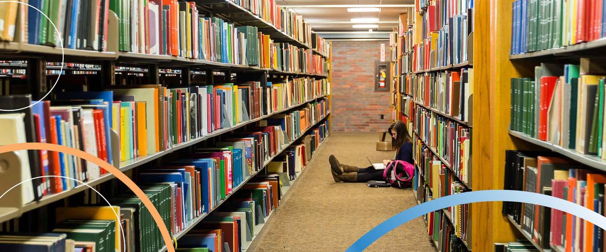 A Boston University student working on a laptop on the floor between library shelves