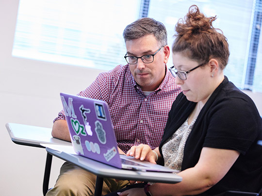 MET Professor Shea Cronin works with a student on a laptop in a classroom