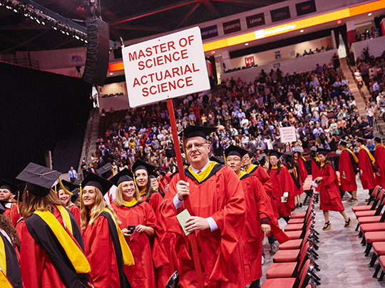 A group of graduates walking into the MET convocation ceremony; a man carrying a sign that reads “Master of Science, Actuarial Science”