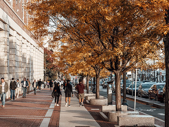 Students walking along a sidewalk with fall foliage next to Agganis Arena