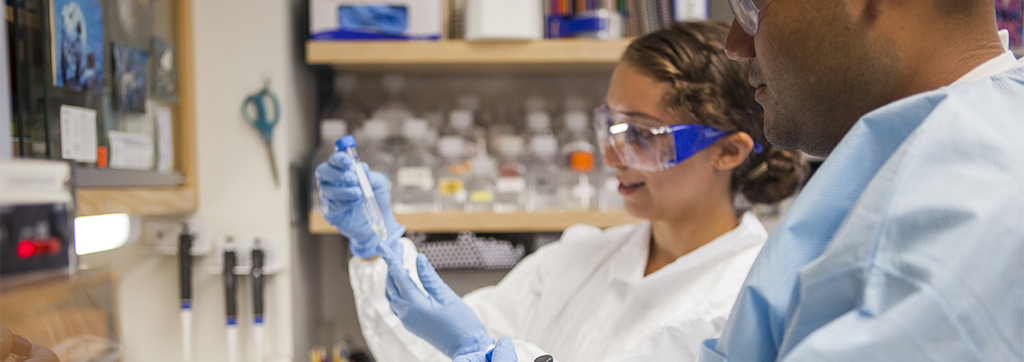 A woman in a laboratory setting wearing safety goggles and a lab coat examines a vial