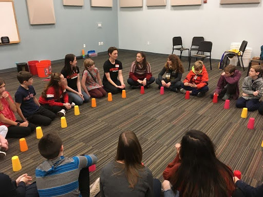Kids with their teacher in a classroom. Everyone is sitting on the floor in a circle, and each person has an upside down cup on the floor in front of them.