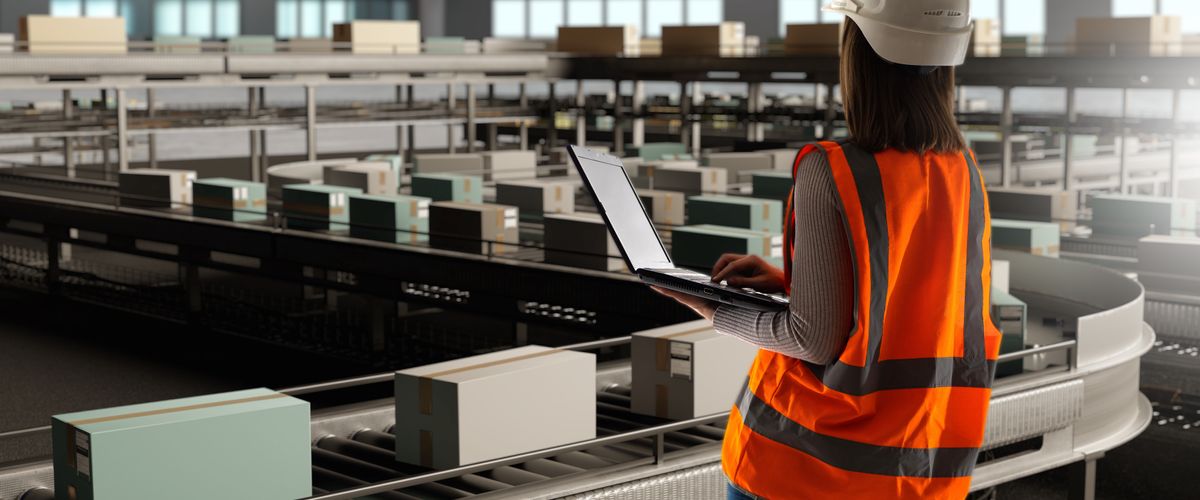 Woman wearing a hardhat and safety vest overlooking a conveyor belt with boxes while holding a laptop