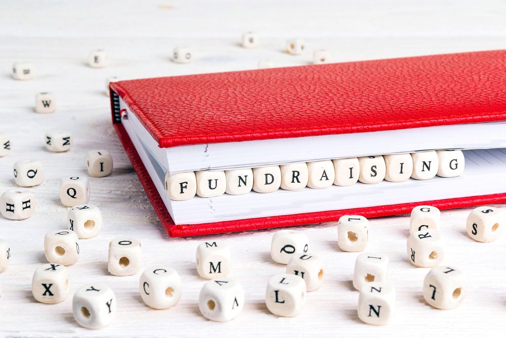 A journal with a red cover is held open slightly by a row of cubic beads spelling the word fundraising. Other cubic alphabet beads are scattered in the foreground and background.