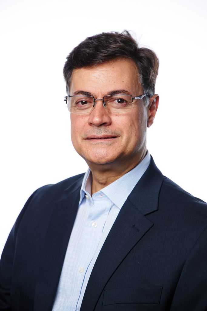 Headshot of a white man with dark hair and glasses. He's looking at the camera and wearing a dark blue suit jacket with a light blue buttonup.