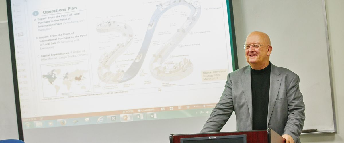 Professor stands in front of a class at a podium in front of a screen showing a graphic