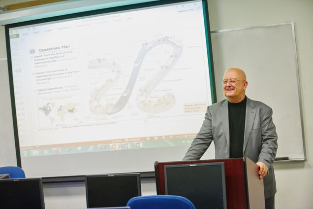 Professors stands in front of a class at a podium in front of a screen showing a graphic