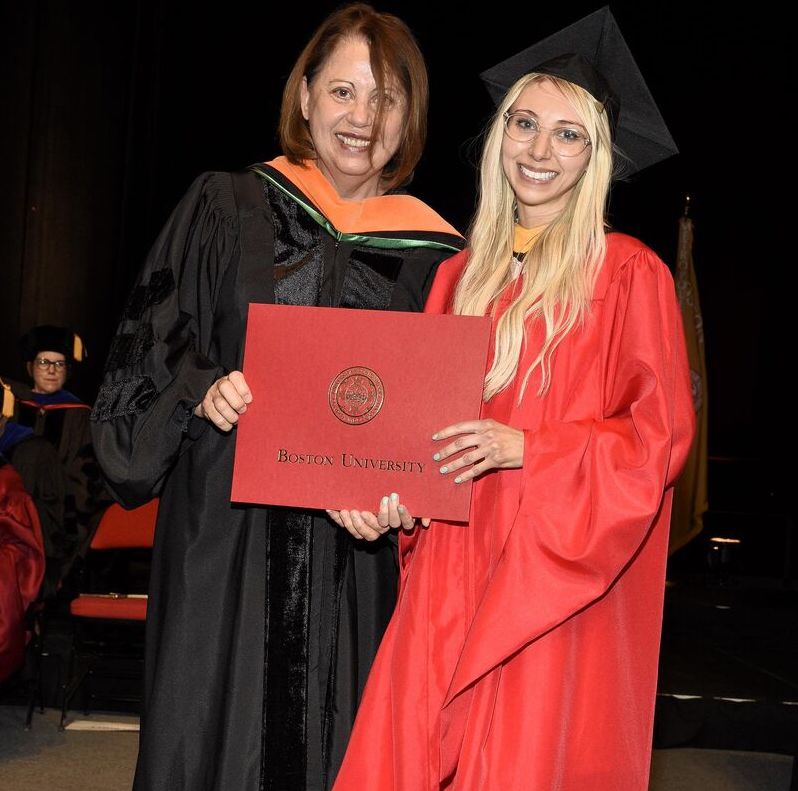 Woman with long blonde hair and glasses in a red robe and black grad cap smiles at the camera as she accepts a diploma from a female faculty member with medium-length brown hair, who is wearing a black robe with a green and orange hood lining.