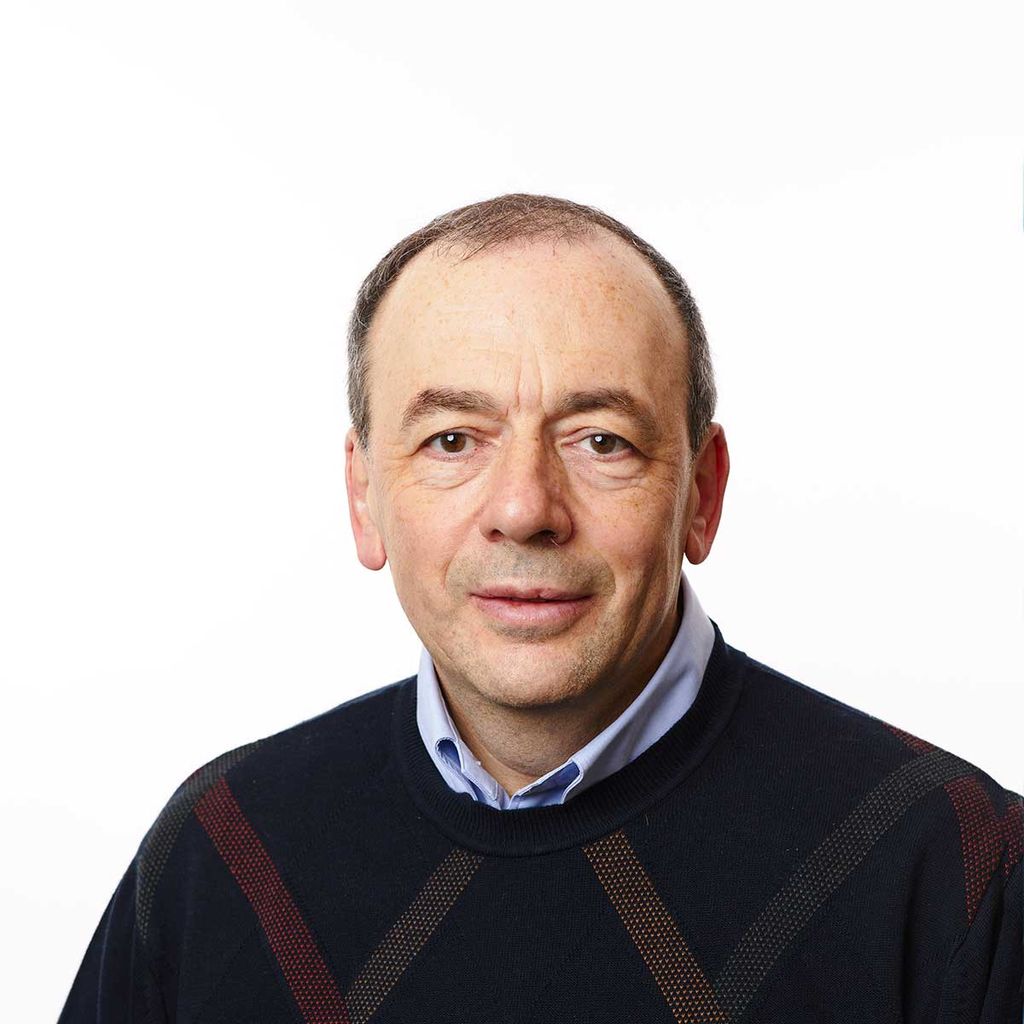 Headshot of an older man looking at the camera. He's wearing a black sweater with a striped pattern over a light blue buttonup.