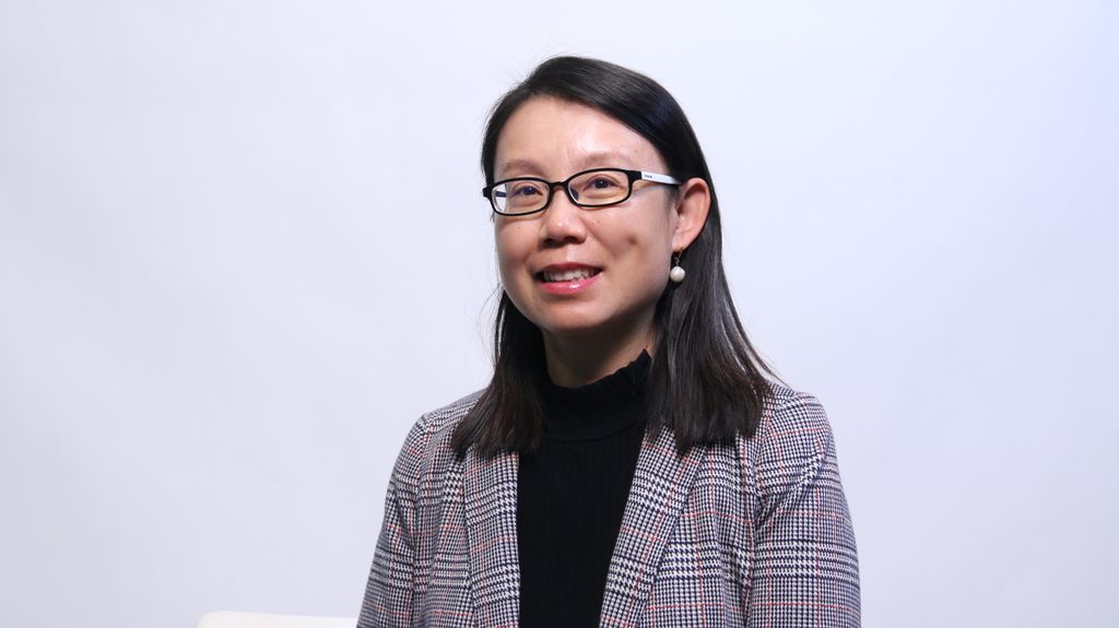 Headshot of a woman with medium-length black hair and glasses smiling at the camera. She's wearing pearl earrings, a plaid striped blazer, and a black shirt.