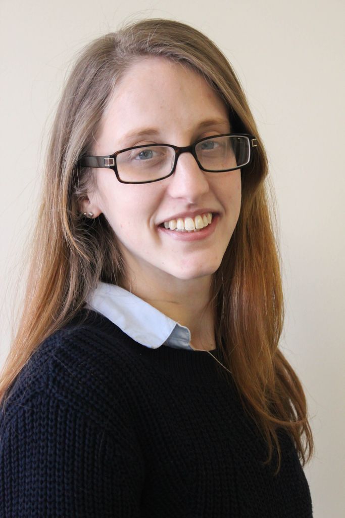 Headshot of a woman with long, dirty-blonde hair and glasses. She's smiling at the camera and wearing a black sweater over a light blue buttonup.