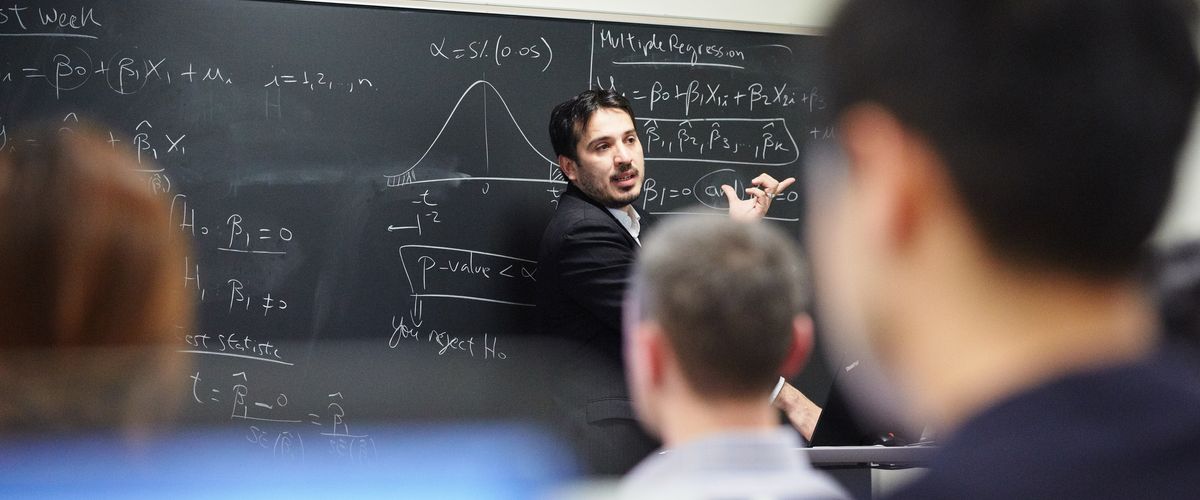 professor stands facing a class in front of a blackboard with equations on it