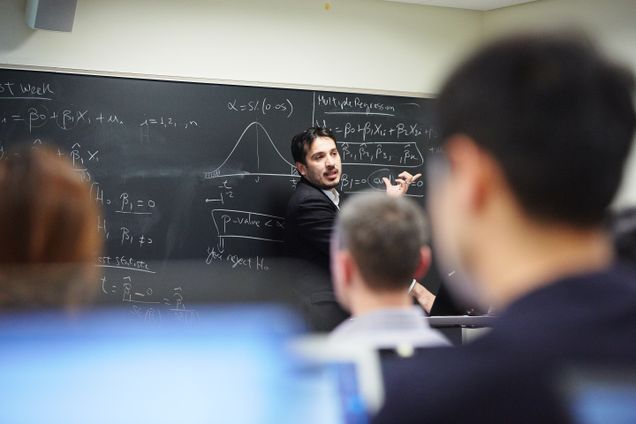 professor stands facing a class in front of a blackboard with equations on it