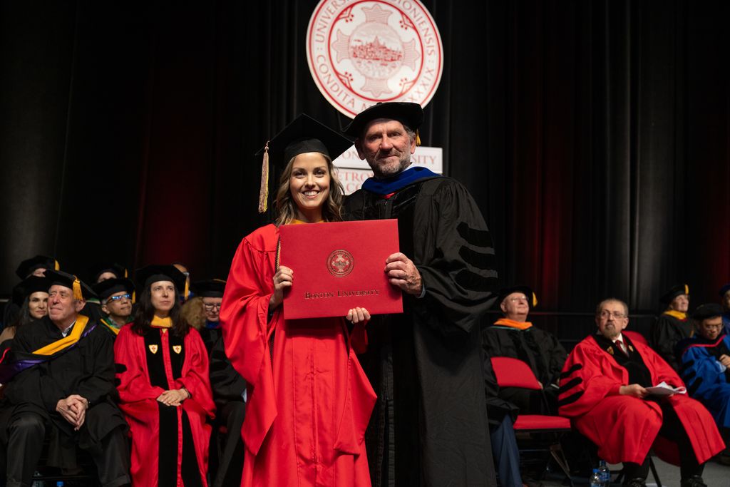 student and professor in graduation regalia hold a diploma between them with other faculty in the background