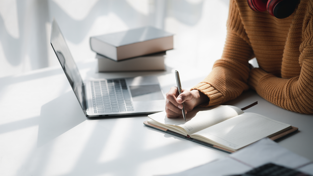 Woman writing in blank journal next to laptop and pile of books