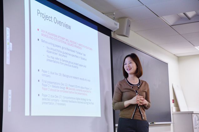 East Asian woman presents at the front of a classroom, looking toward a projecter with a slide reading "project overview."