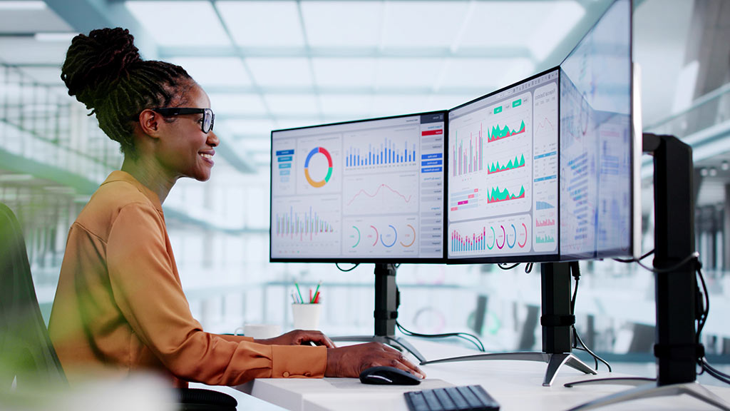 A woman reviews several data charts across three computer monitors in a large office