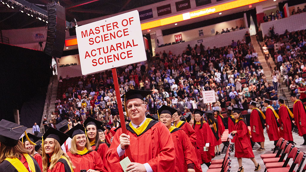 A group of graduates walking into the MET convocation ceremony; a man carrying a sign that reads “Master of Science, Actuarial Science”
