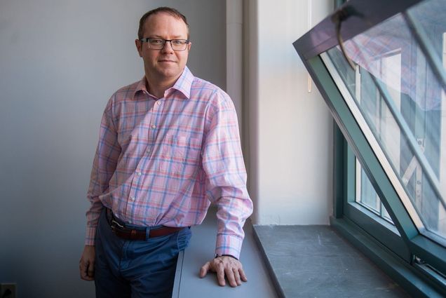 Photo of Benjamin Sovacool. A middle-aged white man wearing glasses and a light pink plaid shirt and jeans poses next to an open window and lightly smiles at the camera.