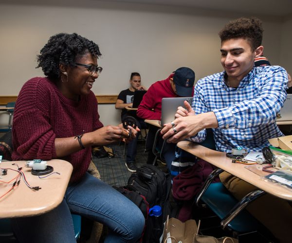 Two smiling students work together in a classroom, helping each other assemble robotics parts.