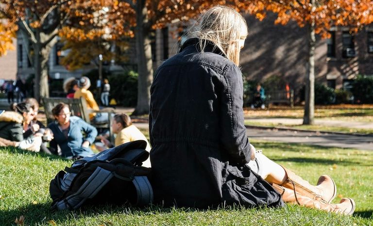 A student sits in the grass and looks at her phone with the colorful fall leaves behind her.