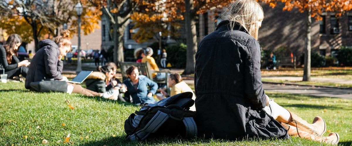 A student sits in the grass and looks at her phone with the colorful fall leaves behind her.