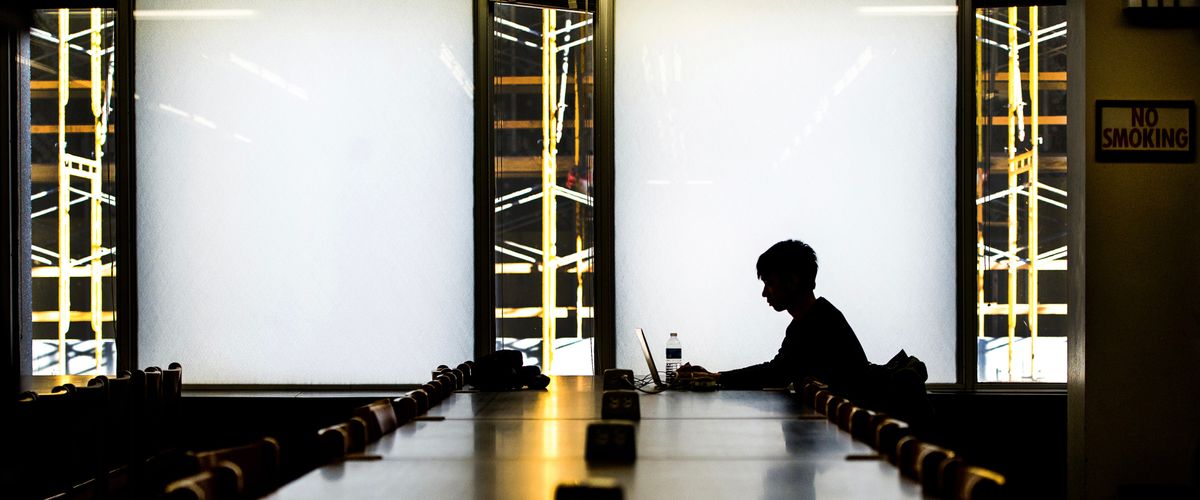 A lone student works on his laptop at the end of a table. He is silhouetted against the windows in Mugar Library.