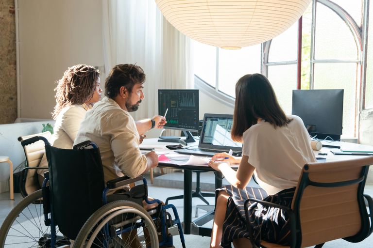 Three coworkers sit together at a desk and review online data, charts, and papers. One man is seated in a wheelchair highlighting the importance diversity and inclusion in the workforce.