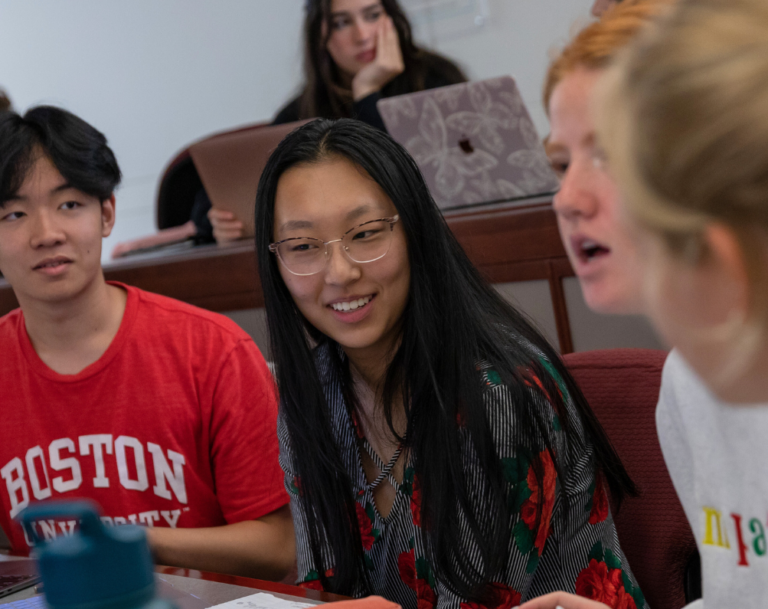 three students engage in the classroom, the one in focus wears a red Boston University T shirt