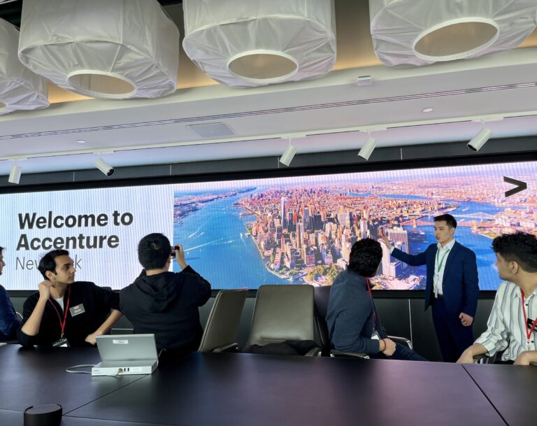 A man standing at the front of a room speaks to seated students at a long conference table. 