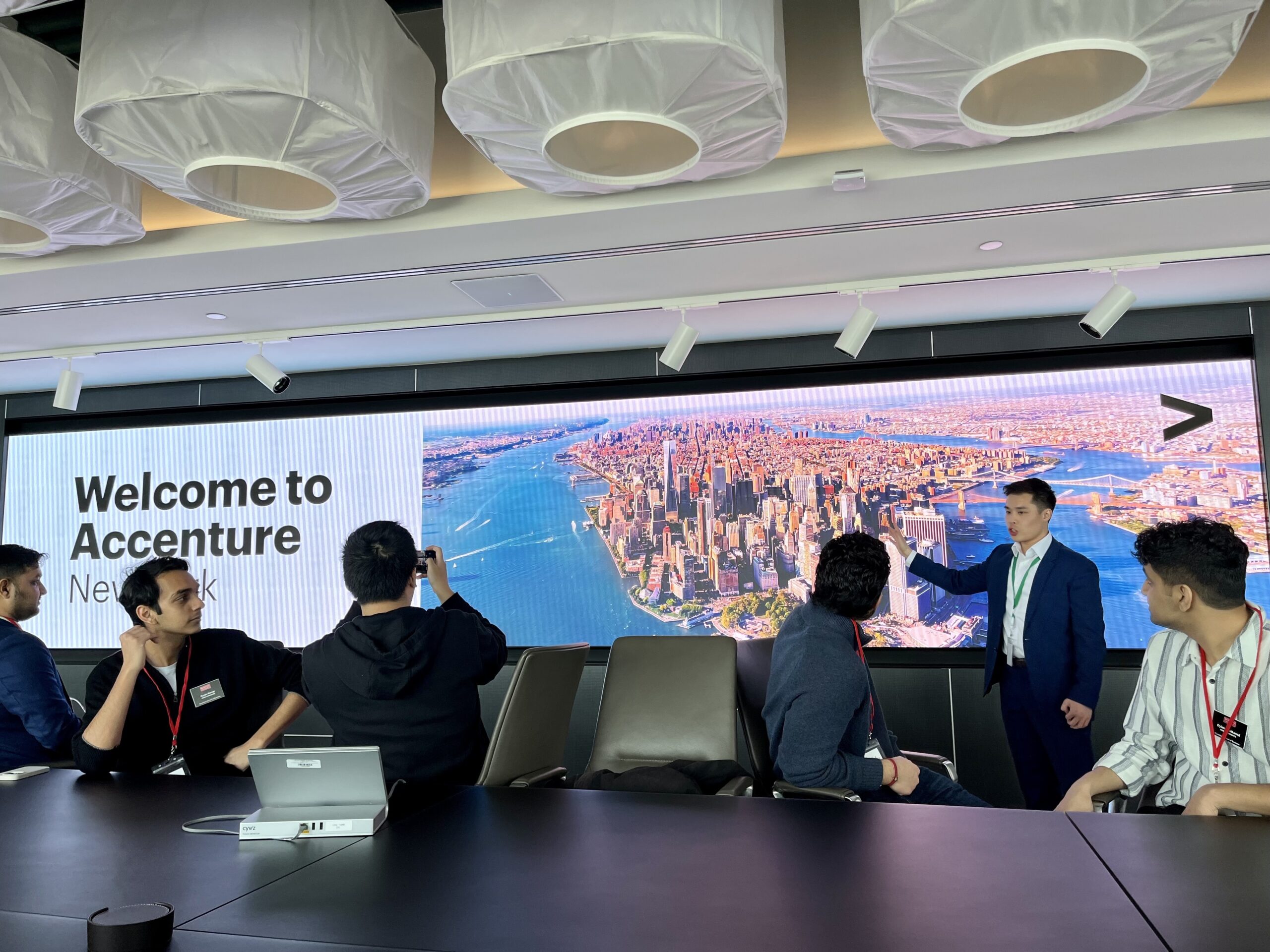 A man standing at the front of a room speaks to seated students at a long conference table. "Welcome to Accenture New York" with an aerial photo of Manhattan is on the display behind him.