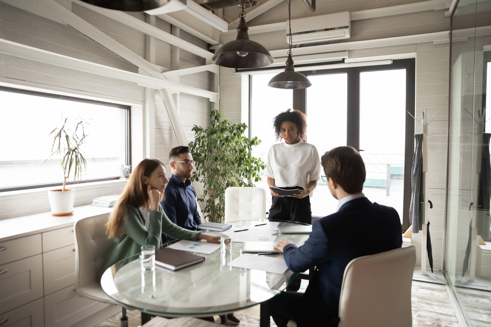 3 business people sit around a table in a modern office building while one person is standing leading the conversation.