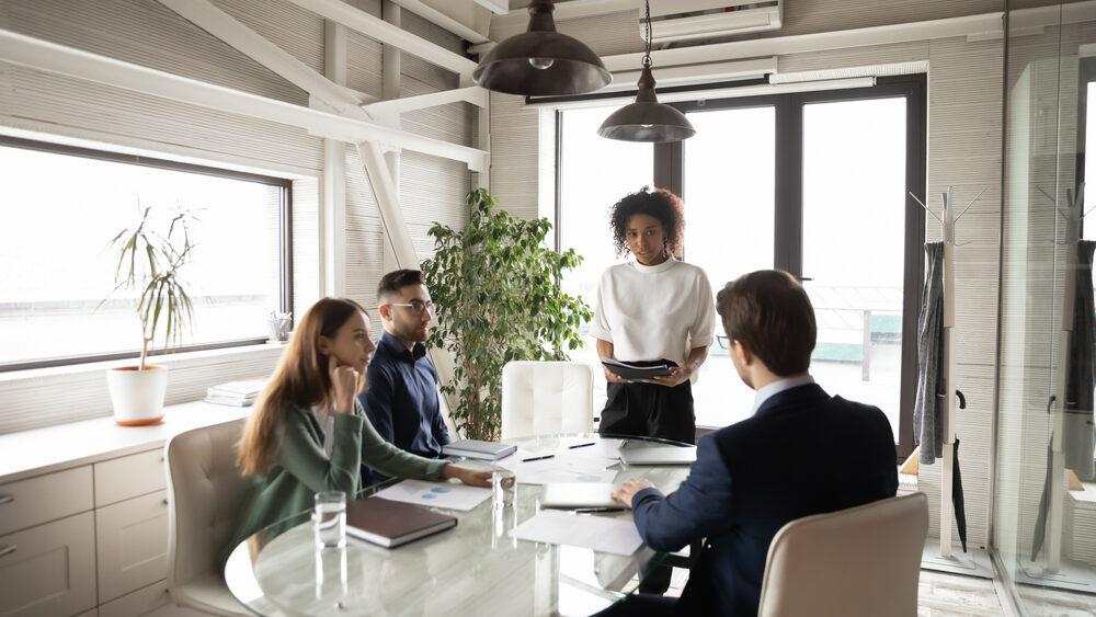 3 business people sit around a table in a modern office building while one person is standing leading the conversation.