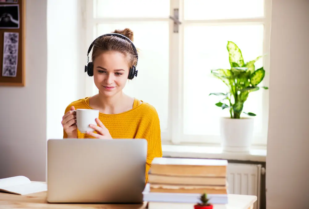 Young women holding coffee cup wearing headset interacting with someone on a laptop.