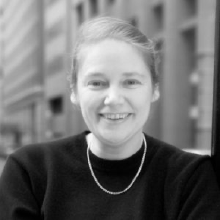 A black and white phot featuring a person with light hair that is pulled up. They are wearing a dark sweater and a necklace.